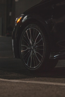 A close-up view of a car tire and wheel, featuring a sleek alloy rim. The surroundings are dimly lit, highlighting the reflective surface of the tire and part of the car body. The foreground is asphalt, suggesting the car is parked or in a driveway.