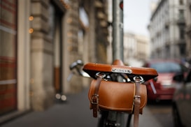 A close-up view of a leather bicycle saddle with an attached brown leather pouch, both branded with the name Brooks. The background is a slightly blurred urban setting with buildings and parked cars, suggesting a street or city environment.