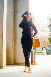 A person wearing a black sweater and trousers stands in a bright outdoor setting, looking to the side while adjusting a graduation cap on their head. The scene includes stone columns, a small wooden podium, and blurred trees in the background, suggesting a formal or academic environment.
