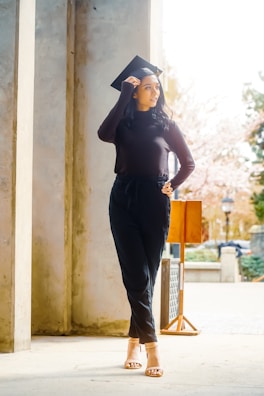 A person wearing a black sweater and trousers stands in a bright outdoor setting, looking to the side while adjusting a graduation cap on their head. The scene includes stone columns, a small wooden podium, and blurred trees in the background, suggesting a formal or academic environment.