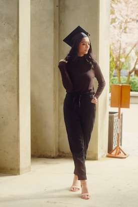 A woman wearing a black graduation cap and a dark outfit stands against a backdrop of concrete columns. She is posing with a subtle smile, with one hand resting on her shoulder. The setting appears to be outdoors with soft lighting, possibly from a nearby tree with pink blossoms in the background.