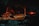 A nighttime scene in a wooded area with a small investigation site. Police tape surrounds a few vehicles, including a vintage car and a police car with flashing lights. A person is adjusting equipment that appears to be set up for illumination or investigation.