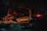 A nighttime scene in a wooded area with a small investigation site. Police tape surrounds a few vehicles, including a vintage car and a police car with flashing lights. A person is adjusting equipment that appears to be set up for illumination or investigation.