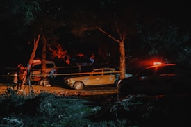 A nighttime scene in a wooded area with a small investigation site. Police tape surrounds a few vehicles, including a vintage car and a police car with flashing lights. A person is adjusting equipment that appears to be set up for illumination or investigation.