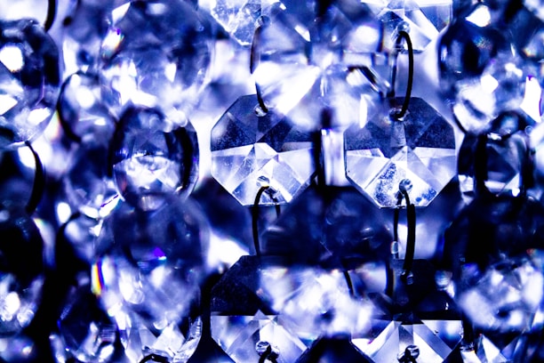 A close-up view of multiple faceted crystal beads or droplets, connected by small metal rings, displaying a vibrant play of light and reflections. The crystals are primarily clear, with hints of blue and purple hues from the lighting.