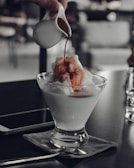 A hand pours a stream of dark liquid, possibly coffee, over a scoop of ice cream in a stylish glass dish. The scene has a modern and minimalistic setting with blurred background elements and a soft focus on the foreground.