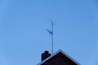 A TV antenna is mounted on the roof of a building with a clear blue sky in the background. The roof features a chimney and has a triangular peak.