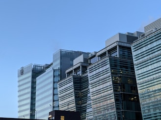 A modern building complex with multiple glass structures, reflecting the clear blue sky. The architecture features clean lines and a contemporary design with several floors visible. Two logos are present near the top of one of the buildings.