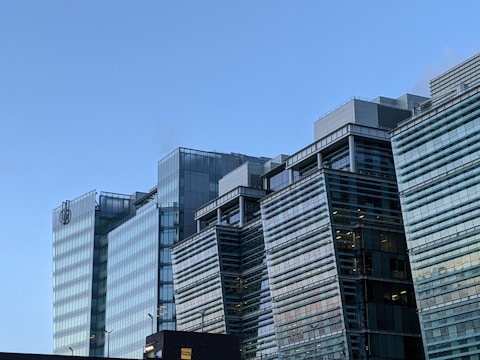 A modern building complex with multiple glass structures, reflecting the clear blue sky. The architecture features clean lines and a contemporary design with several floors visible. Two logos are present near the top of one of the buildings.