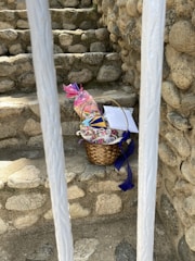 A woven basket containing packaged items and colorful decorations is placed on stone steps surrounded by rock walls. The basket includes a pink-wrapped gift and blue ribbon detailing. Two white bars partially obscure the view.