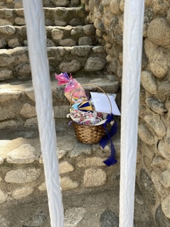 A woven basket containing packaged items and colorful decorations is placed on stone steps surrounded by rock walls. The basket includes a pink-wrapped gift and blue ribbon detailing. Two white bars partially obscure the view.
