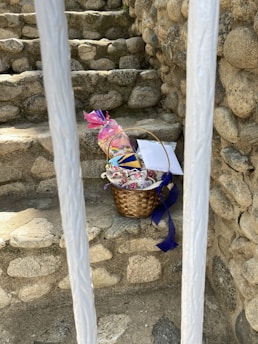 A woven basket containing packaged items and colorful decorations is placed on stone steps surrounded by rock walls. The basket includes a pink-wrapped gift and blue ribbon detailing. Two white bars partially obscure the view.