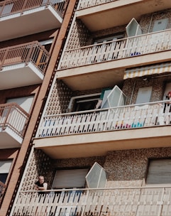 A residential building features several balconies with distinctive patterned walls. The structure is comprised of multiple floors, each with securely closed windows and metal railings. One balcony has an open window, and a person is seen leaning on the railing, partially in shadow.