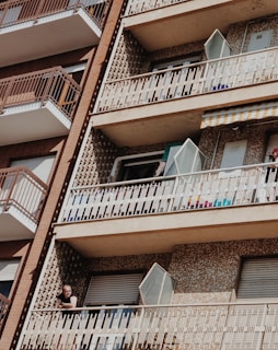 A residential building features several balconies with distinctive patterned walls. The structure is comprised of multiple floors, each with securely closed windows and metal railings. One balcony has an open window, and a person is seen leaning on the railing, partially in shadow.