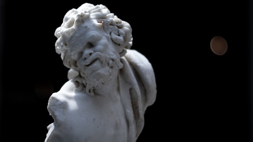 A marble statue of a bearded man with intricate curly hair, partially damaged and set against a dark background. The sculpture's expression appears lively and dynamic, capturing a sense of motion.