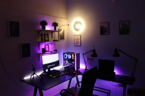 A modern home office setup featuring a dual-monitor desk with a desktop computer. Purple and yellow LED lights illuminate the area, adding a futuristic ambiance. Shelves above the desk hold various items, including potted plants and small tech gadgets. Wall art and motivational quotes adorn the walls, enhancing the creative atmosphere.