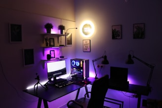 A modern home office setup featuring a dual-monitor desk with a desktop computer. Purple and yellow LED lights illuminate the area, adding a futuristic ambiance. Shelves above the desk hold various items, including potted plants and small tech gadgets. Wall art and motivational quotes adorn the walls, enhancing the creative atmosphere.