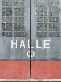 Large industrial doors with weathered glass panes and the word 'HALLE' written across the center. The doors are mostly gray with a red base, displaying signs of wear and age.