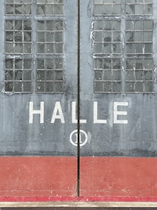Large industrial doors with weathered glass panes and the word 'HALLE' written across the center. The doors are mostly gray with a red base, displaying signs of wear and age.