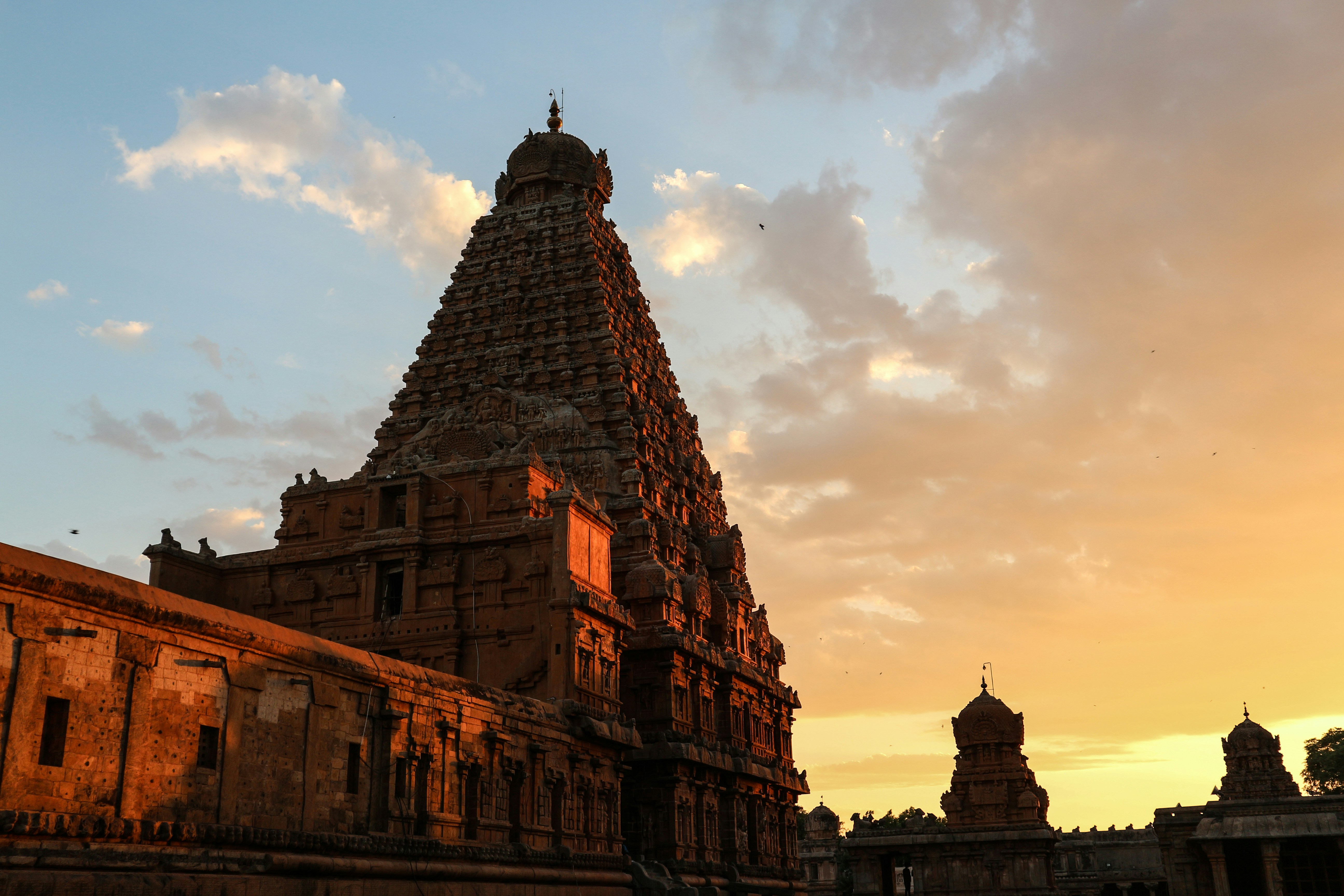 Ancient Shiva temple exterior at dusk, warm amber floodlights illuminating carved stone facade, deep indigo sky, sacred atmosphere, dramatic shadows