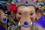A detailed depiction of a Ganesha statue adorned with intricate jewelry and a decorative crown, featuring vibrant blue gemstones surrounded by gold and diamonds. The statue has a serene expression, with traditional Indian symbols painted on its forehead and trunk. Rich textures and colors create a visually captivating effect.