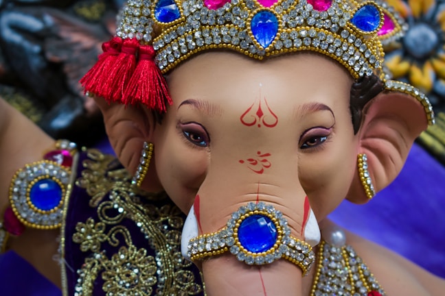 A detailed depiction of a Ganesha statue adorned with intricate jewelry and a decorative crown, featuring vibrant blue gemstones surrounded by gold and diamonds. The statue has a serene expression, with traditional Indian symbols painted on its forehead and trunk. Rich textures and colors create a visually captivating effect.