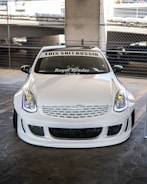 A customized white car is parked in a garage with a prominent front bumper and grill design. The windshield displays text decals reading 'THIS SHIT BUSSIN' and 'Royal Spades'. The background includes concrete pillars and a chain-link fence.