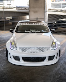 A customized white car is parked in a garage with a prominent front bumper and grill design. The windshield displays text decals reading 'THIS SHIT BUSSIN' and 'Royal Spades'. The background includes concrete pillars and a chain-link fence.