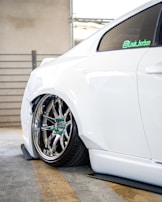 The image features the rear side of a white car with custom chrome alloy wheels. The car is parked in a garage, indicated by the concrete floor and metal railing in the background. The suspension is notably lowered, causing the wheel to sit very close to the wheel arch.