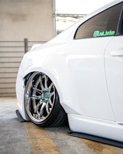 The image features the rear side of a white car with custom chrome alloy wheels. The car is parked in a garage, indicated by the concrete floor and metal railing in the background. The suspension is notably lowered, causing the wheel to sit very close to the wheel arch.