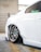 The image features the rear side of a white car with custom chrome alloy wheels. The car is parked in a garage, indicated by the concrete floor and metal railing in the background. The suspension is notably lowered, causing the wheel to sit very close to the wheel arch.