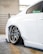 The image features the rear side of a white car with custom chrome alloy wheels. The car is parked in a garage, indicated by the concrete floor and metal railing in the background. The suspension is notably lowered, causing the wheel to sit very close to the wheel arch.