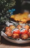 A bowl filled with a collection of decorated eggs, some of which are wrapped in Kinder Surprise wrappers. The background includes leafy green plants and a soft focus on what appears to be a baked dish.