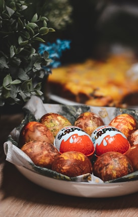 A bowl filled with a collection of decorated eggs, some of which are wrapped in Kinder Surprise wrappers. The background includes leafy green plants and a soft focus on what appears to be a baked dish.
