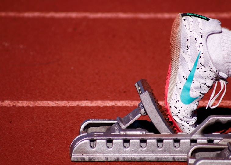 A close-up of a white athletic shoe with colorful speckles and a blue swoosh logo, resting on a starting block on a red track field. The shoe is positioned as if ready for a race, with a visible foot wearing a white sock inside the shoe.