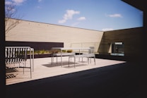 A modern outdoor terrace with minimalist furniture, including metal benches and a table. The area is surrounded by wooden panel walls and features a clear blue sky with a few clouds visible. Sunlight casts distinct shadows on the wooden floor, creating a serene and open atmosphere.