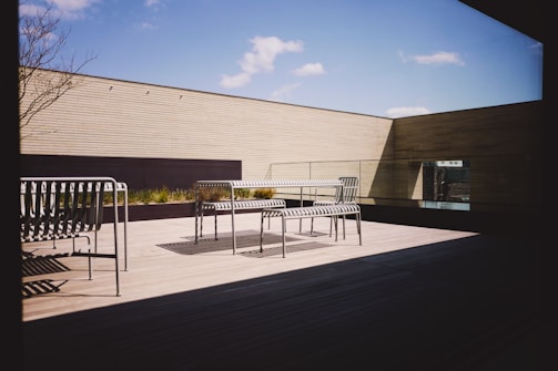 A modern outdoor terrace with minimalist furniture, including metal benches and a table. The area is surrounded by wooden panel walls and features a clear blue sky with a few clouds visible. Sunlight casts distinct shadows on the wooden floor, creating a serene and open atmosphere.