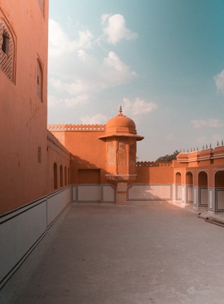 A spacious courtyard with orange walls featuring traditional architecture. The area is bordered by intricate latticework and arched doorways, with a central dome-like structure. Soft light casts gentle shadows, creating a serene and peaceful atmosphere.