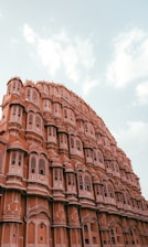 A majestic architectural structure with intricate lattice windows and a series of small balconies. The building features a distinctive pink hue, typical of the iconic Hawa Mahal in Jaipur, India. The sky is partly cloudy, adding to the grandeur of the scene.