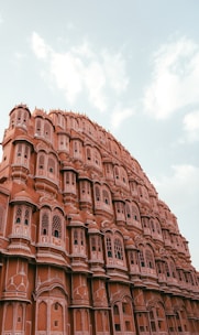 A majestic architectural structure with intricate lattice windows and a series of small balconies. The building features a distinctive pink hue, typical of the iconic Hawa Mahal in Jaipur, India. The sky is partly cloudy, adding to the grandeur of the scene.