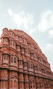 A majestic architectural structure with intricate lattice windows and a series of small balconies. The building features a distinctive pink hue, typical of the iconic Hawa Mahal in Jaipur, India. The sky is partly cloudy, adding to the grandeur of the scene.
