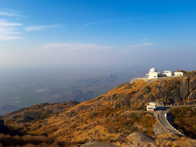 An observatory with a white dome sits atop a rocky, dry hill covered in brown and yellow vegetation. A narrow winding road leads up to the observatory. The sky is clear with a gradient of blue, and there is a vast, hazy landscape stretching into the distance.