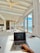 A person is using a laptop on a bed in a bright, airy room with large windows overlooking the ocean. The wooden ceiling features exposed beams and a ceiling fan. The room is furnished in a minimalist style with light-colored decor, providing a relaxing and open atmosphere. A small set of items, including a pen and a watch, are placed nearby on the bed.