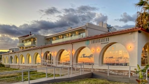 A two-story building with arched windows and a restaurant sign is situated against a cloudy sky. The setting sun casts a warm glow on the structure, which features a modern design with balconies and outdoor seating. A palm tree is visible on the right side, adding to a coastal ambiance.