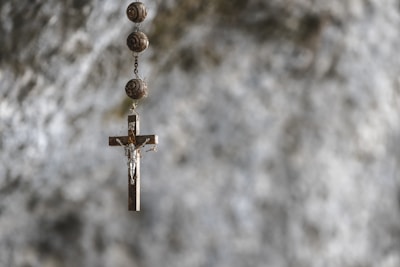 A wooden cross with a metal depiction of Jesus hangs from a chain adorned with spherical beads. The background is blurred, featuring a textured stone or concrete surface in shades of gray.