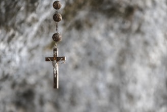 A wooden cross with a metal depiction of Jesus hangs from a chain adorned with spherical beads. The background is blurred, featuring a textured stone or concrete surface in shades of gray.