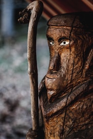 A detailed wooden sculpture depicts a figure with a staff, showcasing intricate carvings and texture. The eyes are prominent, adding a lifelike quality to the figure. The background is blurred, drawing attention to the sculpture.