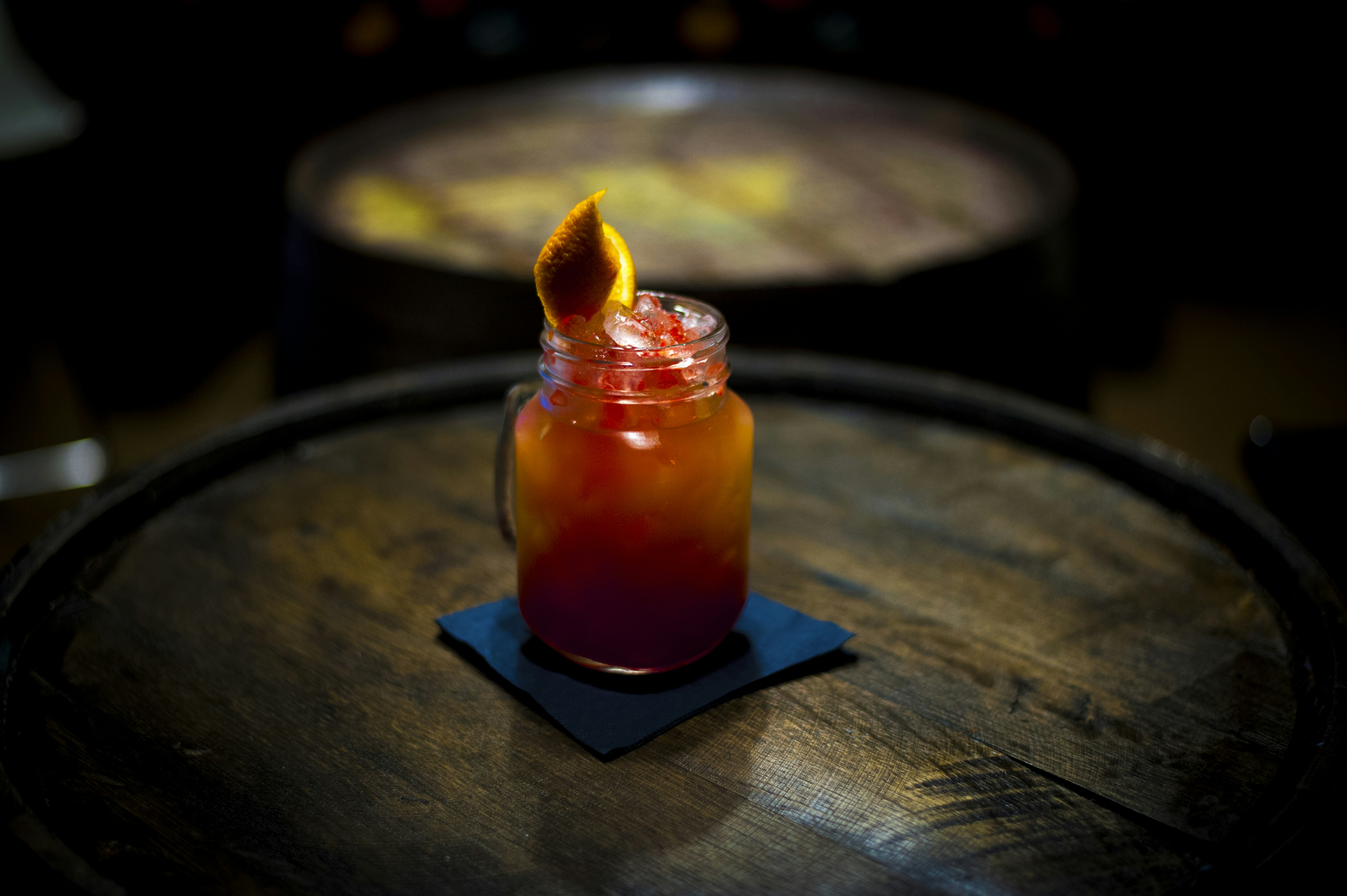 Deep red Negroni-style cocktail in rocks glass with charred orange peel, dark wood bar surface