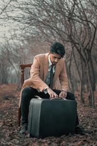 A man in a beige overcoat and black pants sits on a wooden chair in a leaf-covered forest. He is leaning forward, focusing on a large black suitcase placed in front of him.