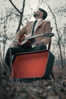 A man in a tan blazer and tie is sitting on a chair outdoors, holding a classical guitar. An open, empty guitar case lies on the ground nearby. The barren trees and dry leaves suggest a winter or autumn setting.